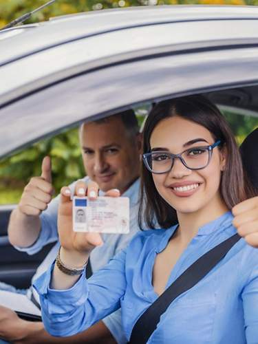 Eine Fahranfängerin hält Ihren Führerschein hoch und freut sich. Ihr Fahrlehrer sitzt daneben.